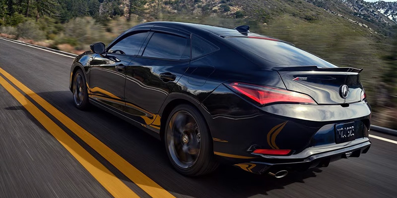 A sleek black 2025 Acura Integra driving on a highway with yellow lines, surrounded by trees and mountains.