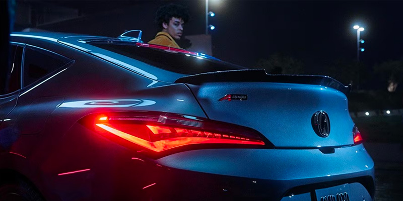 A sleek blue 2025 Acura Integra car with bright red taillights, parked at night with a person in the background.