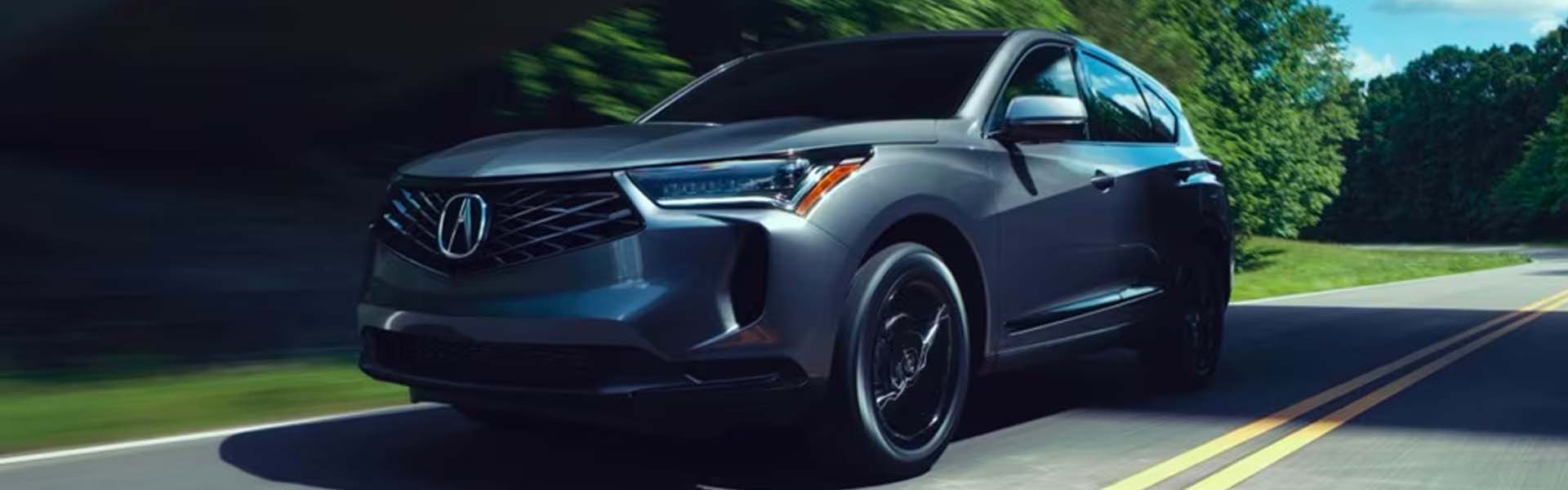 A sleek gray 2025 Acura RDX SUV driving on a winding road surrounded by green trees.