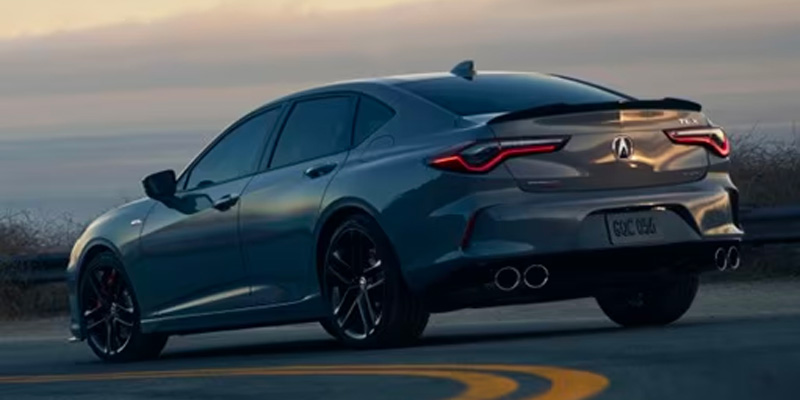 A sleek gray 2025 Acura TLX parked on a curvy road at sunset, featuring stylish taillights and dual exhaust.