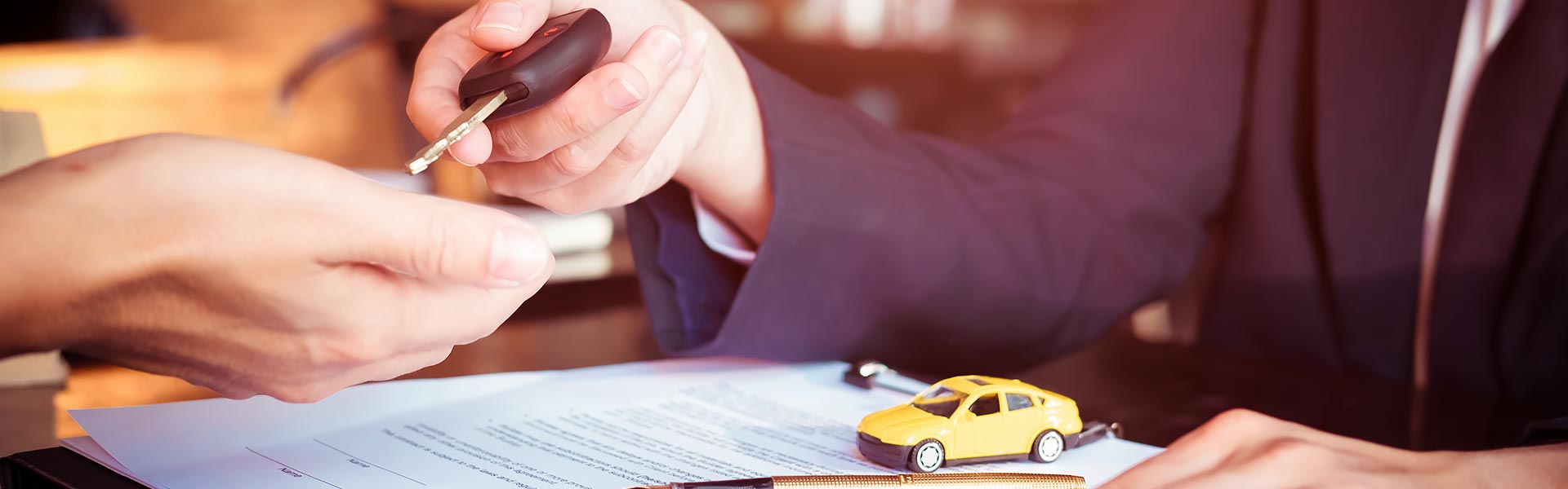A person hands over car keys to another person, with a toy car and documents on the table.