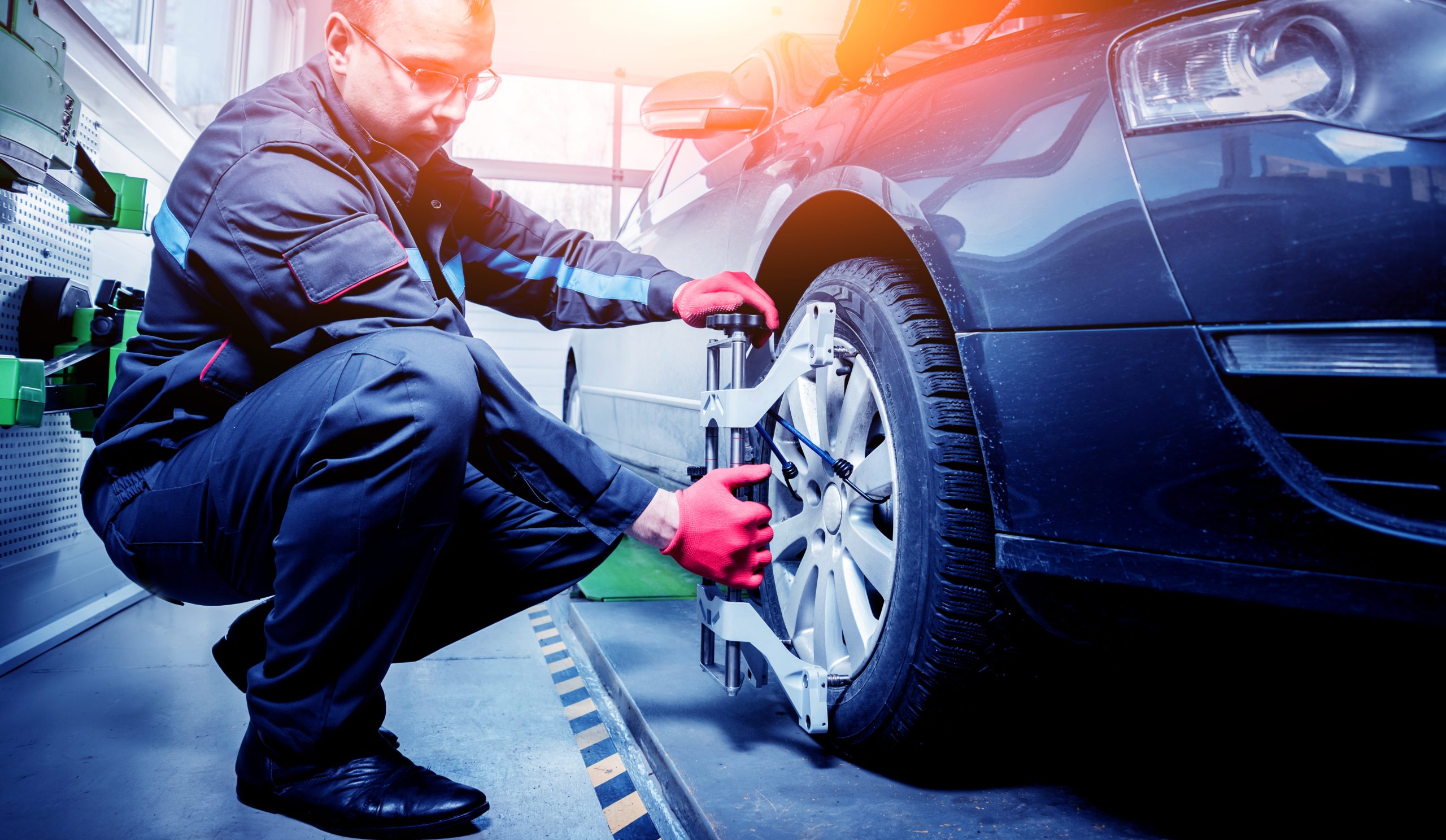 A mechanic in gloves measures a car's wheel alignment in a garage.